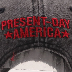 Macro detail of red 'PRESENT-DAY AMERICA' embroidery on dark grey washed cotton baseball cap