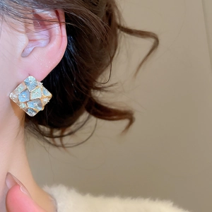 Detail showing gold-plated square stud earrings with blue floral mosaic pattern on ear