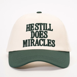 Two-tone beige and dark green baseball cap with HE STILL DOES MIRACLES embroidery