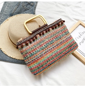 Detail of colorful woven straw bag featuring golden square handles and brown pom-poms
