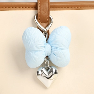 Light blue fabric bow keychain with silver heart pendant on a beige leather bag