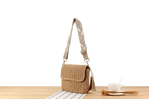 Detail of hand-woven straw crossbody bag with striped strap and tassel on wooden table