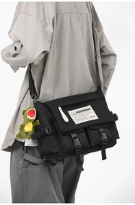 Detail of black utility messenger bag with a green dinosaur plush charm on a model