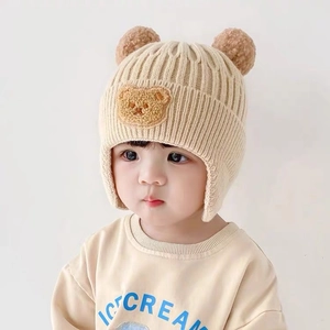 Detail of beige knit baby hat with ear flaps, bear embroidery and brown faux fur pom poms