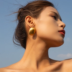 Macro view of matte 18k gold oval earrings worn by a model against a blue sky