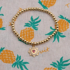 Detail of gold beaded bracelet with heart charm and red zircon on a textured background