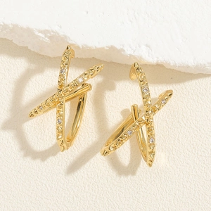 Detail of gold X-shape earrings with zircon studs on a textured stone background