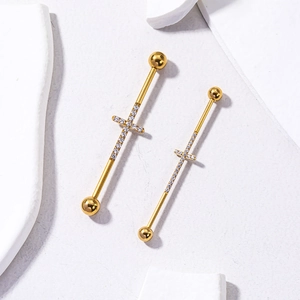 Détail de deux piercings industriels dorés avec pendentifs croix en zircon sur pierre texturée