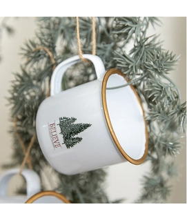 Close-up of white enamel Christmas tree mug with gold rim and Believe text hanging on a pine branch