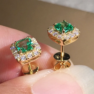 Macro view of gold halo stud earrings with emerald-cut green zircon held between fingers