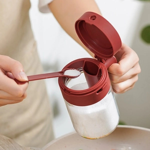 Kitchen glass seasoning jar with dark red flip lid and spoon showing salt dispensing