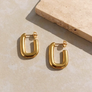 Détail de boucles d'oreilles en forme de U dorées en acier sur un fond de pierre sobre