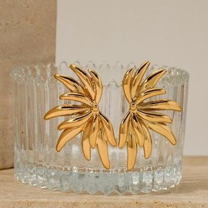Golden feather-shaped stainless steel earrings on a glass edge with soft natural lighting