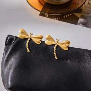 Boucles d'oreilles libellule en acier doré mat présentées sur un sac en cuir noir chic