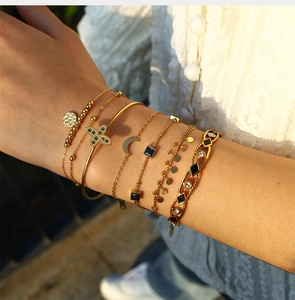 Détail de bracelets dorés avec breloques lune, croix et pierres carrées noires en couches