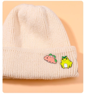 Detail of cute dinosaur and frog enamel pins on a beige knitted beanie hat surface