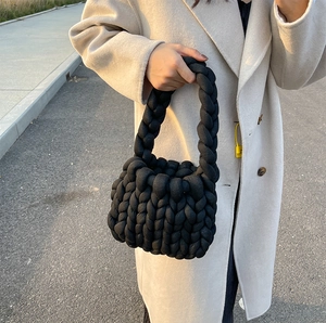 Close-up of a chunky black knitted handbag held by a model in a beige coat outdoors