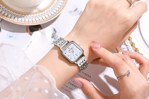 Gros plan d'une montre carrée en nacre avec bracelet argenté porté avec de la dentelle