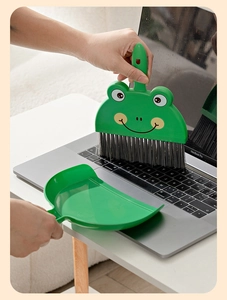 Mini green frog broom set cleaning a laptop keyboard on a wooden desk top view