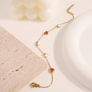 Dettaglio del bracciale a cuore dorato con zirconi blu e rosa su uno sfondo crema