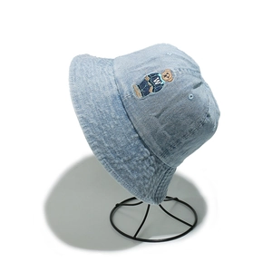 Close-up of light blue denim bucket hat with detailed bear embroidery on a metal stand