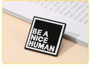 Close-up of square enamel pin with 'BE A NICE HUMAN' text on a neutral background