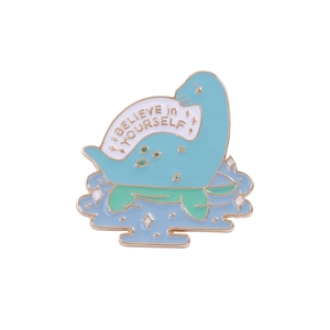 Close-up of cute enamel dinosaur brooch with 'BELIEVE in YOURSELF' text and gold edges
