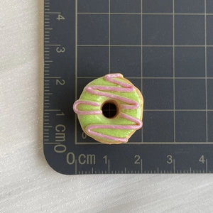 Macro detail of a 2cm green resin donut charm with pink icing on a measurement mat