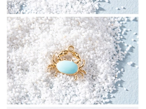 Macro shot of light blue enamel crab pendant with gold-plated edges on white sand