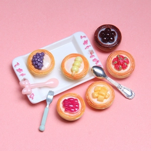 Miniature artificial fruit tarts set on a pink background with tiny dessert utensils