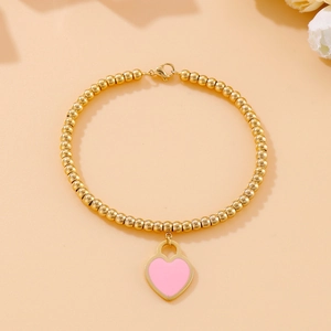 Detail of beaded 18k gold plated bracelet with a pink enamel heart charm on a peach surface