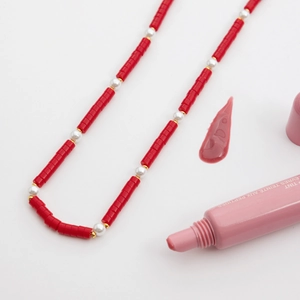 Detail of red stone and pearl necklace next to a pink peptide lip tint on white background