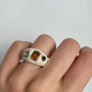 Close-up of a wide cream resin ring featuring amber, green, and clear stones on a finger