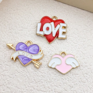 Three enamel heart and wing charms with LOVE lettering on a textured beige surface
