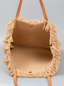Beige woven straw tote bag with fringe details and brown leather handles on grey background