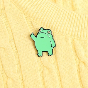 Detailed view of a green funny frog enamel pin on a yellow knitted sweater surface