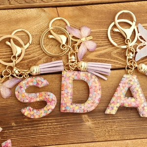 Pink resin alphabet keychains with glitter butterfly and purple tassel on wood surface