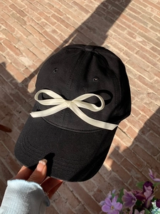 Detail of a black baseball cap featuring a large white satin bow against a brick wall