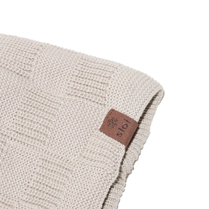 Dettaglio macro del berretto in maglia beige con etichetta in pelle marrone e fiocco