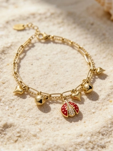 Détail d'un bracelet plaqué or 18k avec breloque coccinelle rouge et cœurs sur sable blanc