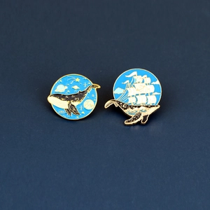 Close-up of enamel whale pins featuring starry sky and sailing ship designs with gold trim