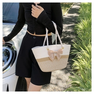 Detail of a woven straw tote bag with a beige ribbon bow and rope handle in outdoor setting