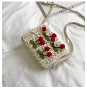 Detail of straw crossbody bag featuring red plush cherry accents and thick cotton rope strap