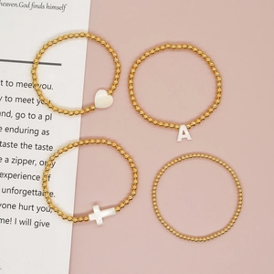 Beaded gold bracelets with heart, cross, and letter A shell charms on pink background