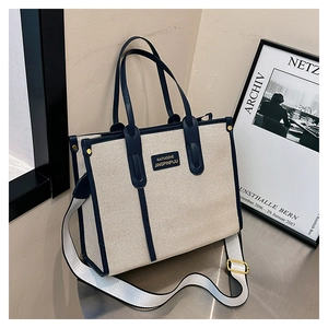 Detail of canvas tote bag showing beige fabric, dark blue leather trim, and logo patch