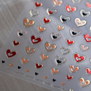 3D heart nail art stickers with red, gold glitter and metallic outlines on textured grey fabric