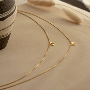Detail of two gold box chain necklaces with letter B and N pendants on beige backdrop