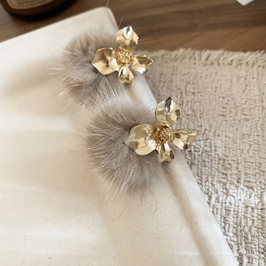 Close-up of golden floral stud earrings with plush grey-brown pompom pendants