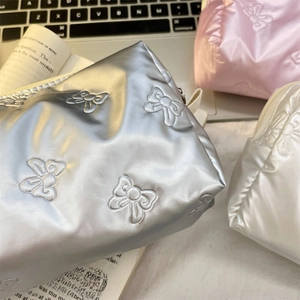Three shiny bow-patterned makeup bags in silver, pink, and white on a lifestyle desk setup