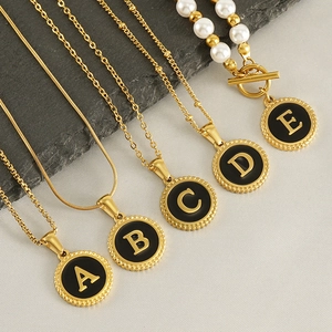 Five gold necklaces with black initial pendants A to E on stone and fabric background